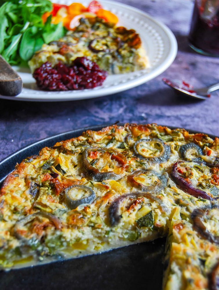 A photo of a crustless quiche with peppers and red onion rings on top. A white plate with a slice of the quiche,salad and chutney can be seen in the background.