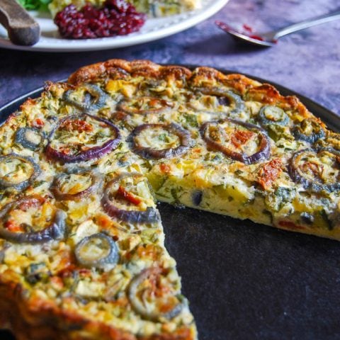 A black plate with a crustless quiche. A cut piece of the quiche with salad on a plate can be seen in the background.