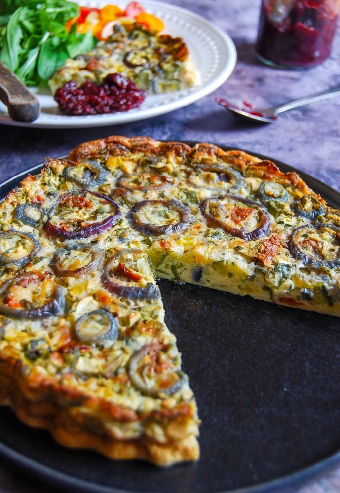 A Photo of a crustless quiche. A slice of cut quiche with salad can be seen in the background on a white plate.