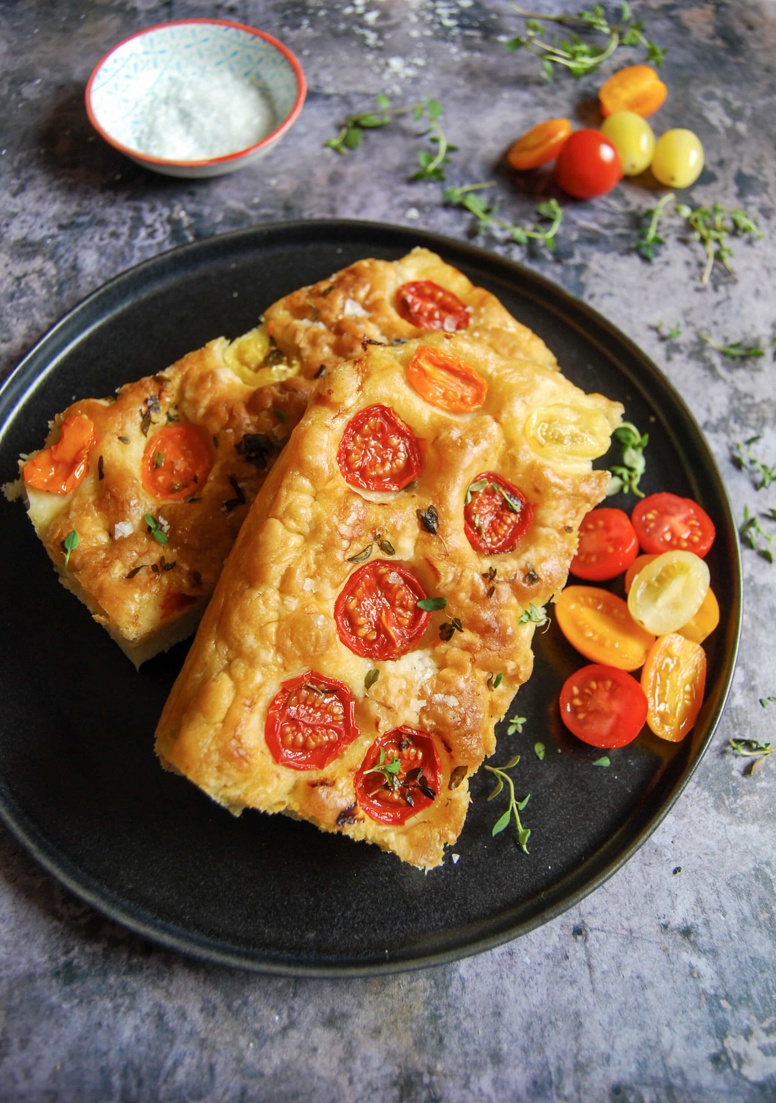 Slices of Focaccia bread with cherry tomatoes and thyme on a black plate. 