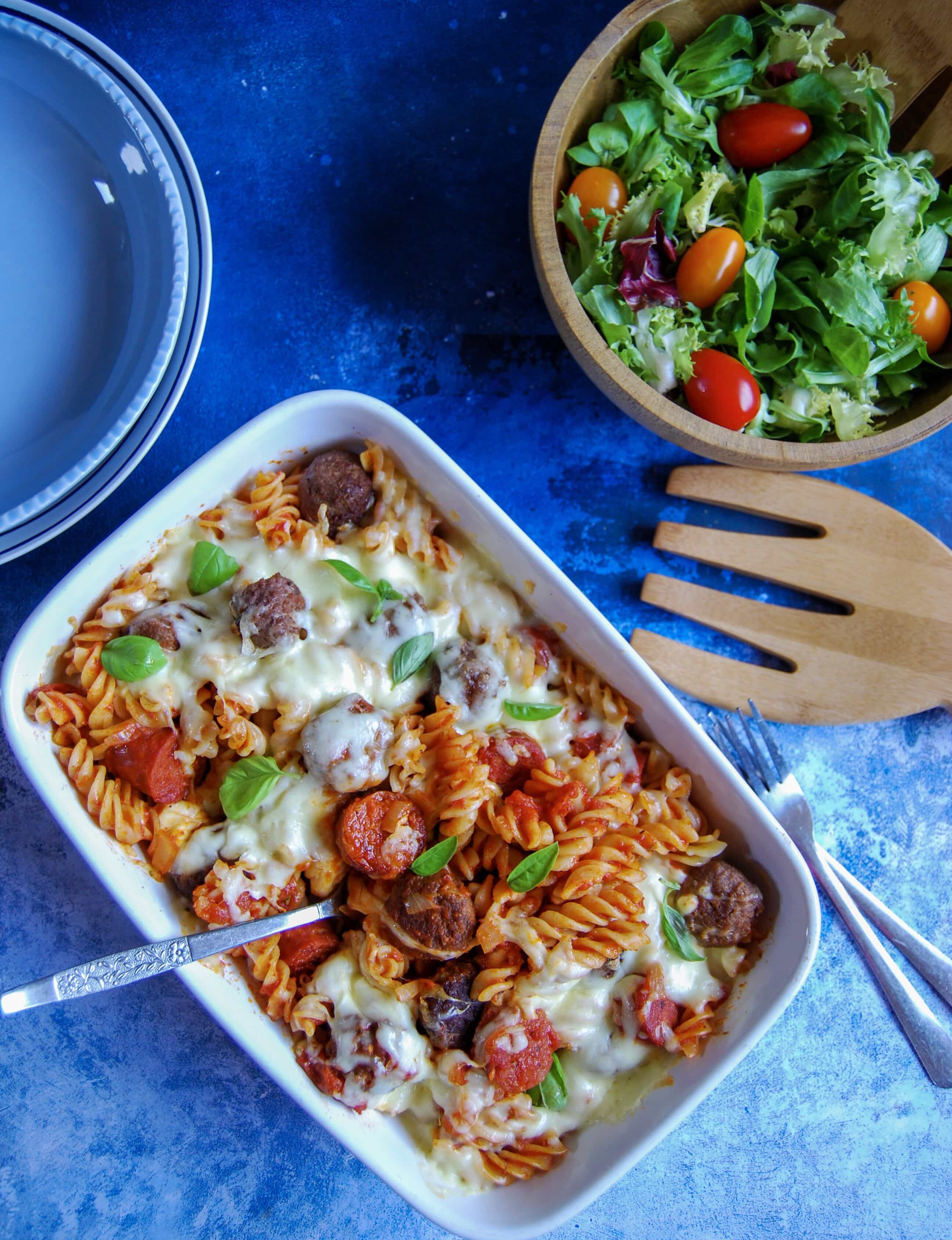 A flatlay photo of a meatball mozzarella pasta bake on a blue background. A bowl of salad and salad server can be seen top right.