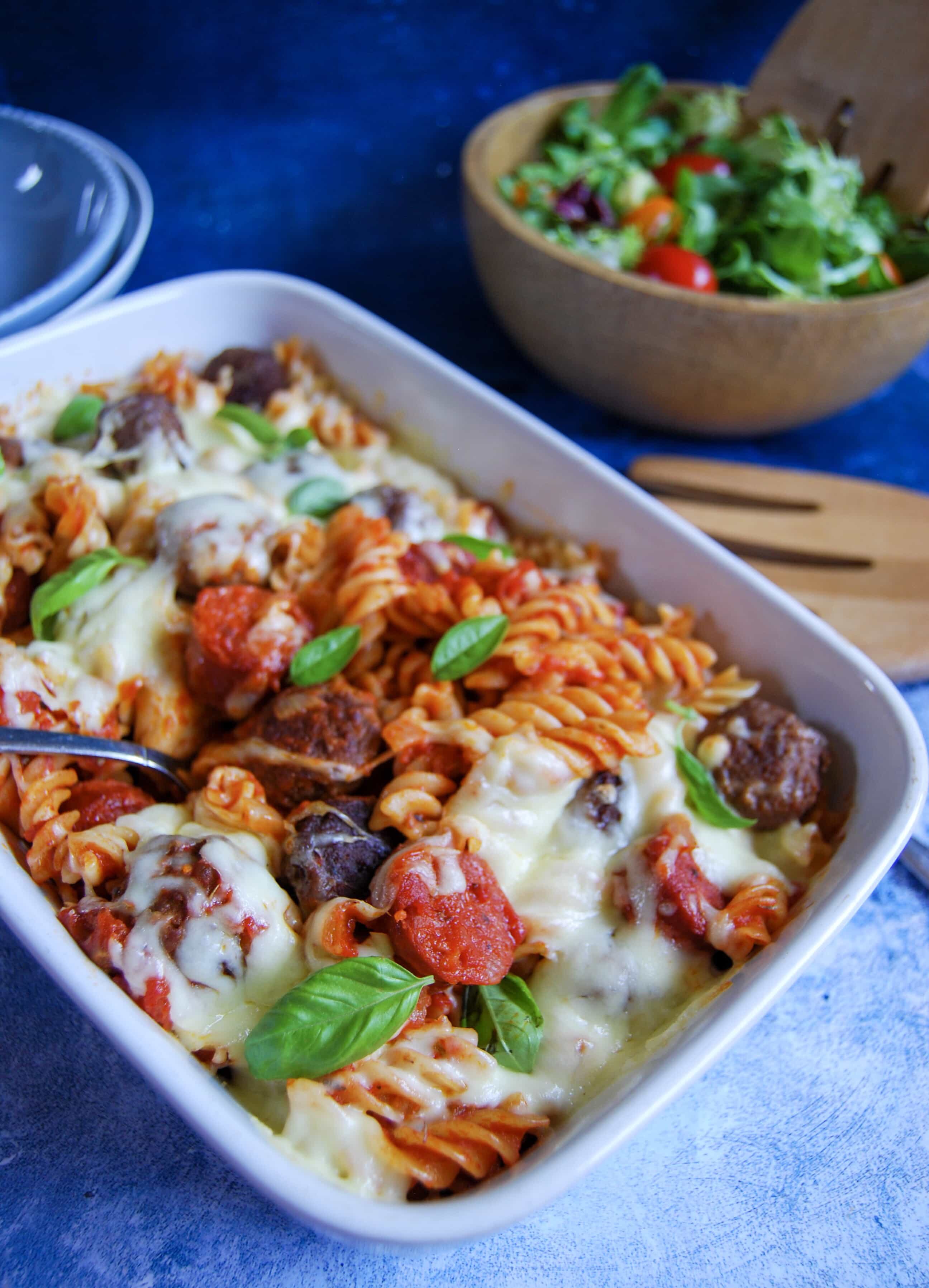 A picture of a mozzarella meatball pasta bake. A wooden bowl of salad can be seen in the background.