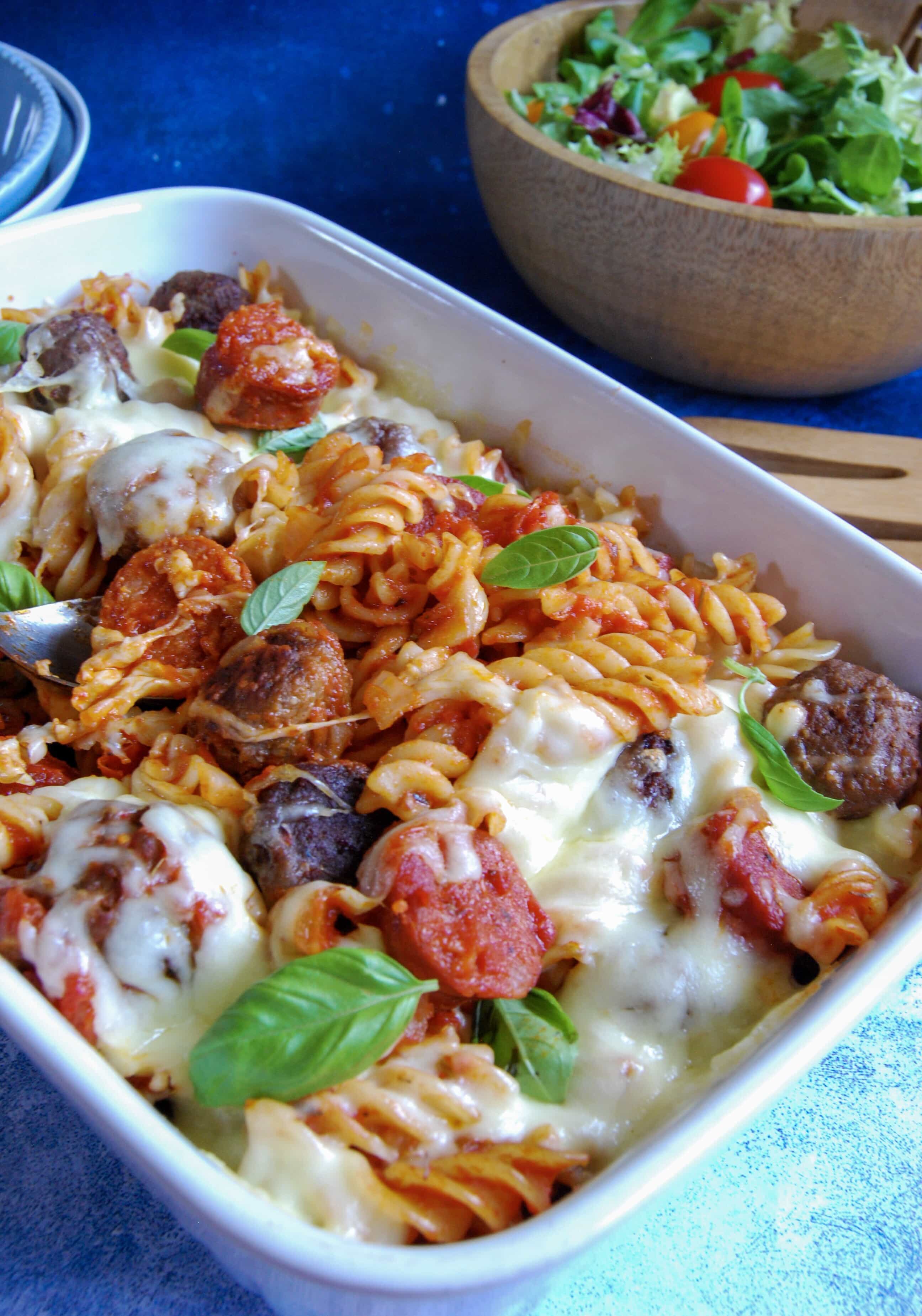 A picture of a mozzarella meatball pasta bake. A wooden bowl of salad can be seen in the background.