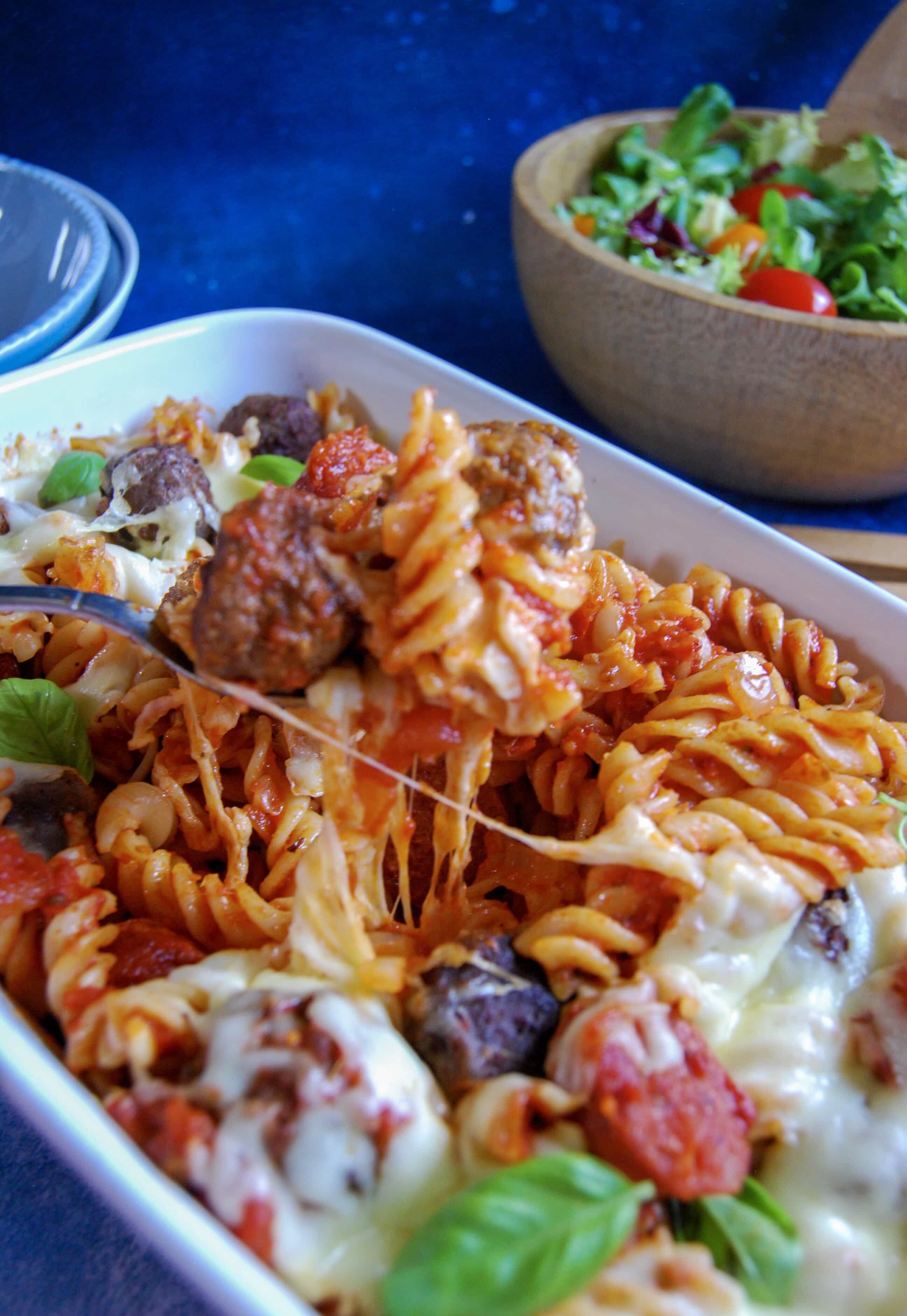 A picture of a mozzarella meatball pasta bake with gooey cheese. A wooden bowl of salad can be seen in the background.