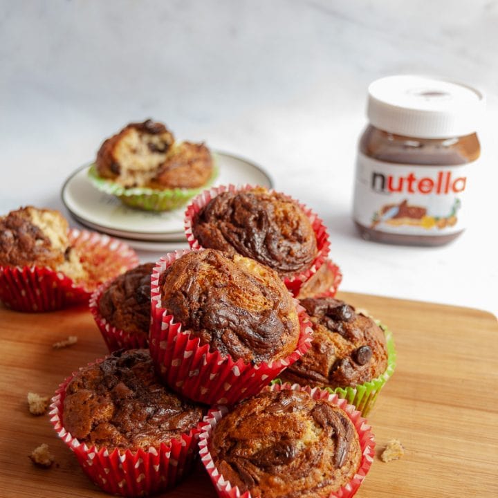 chocolate chip banana nutella mchocolate banana muffins in red and green muffin liners on a wooden board. In the background is a plate with a torn open muffin beside a jar of Nutella.