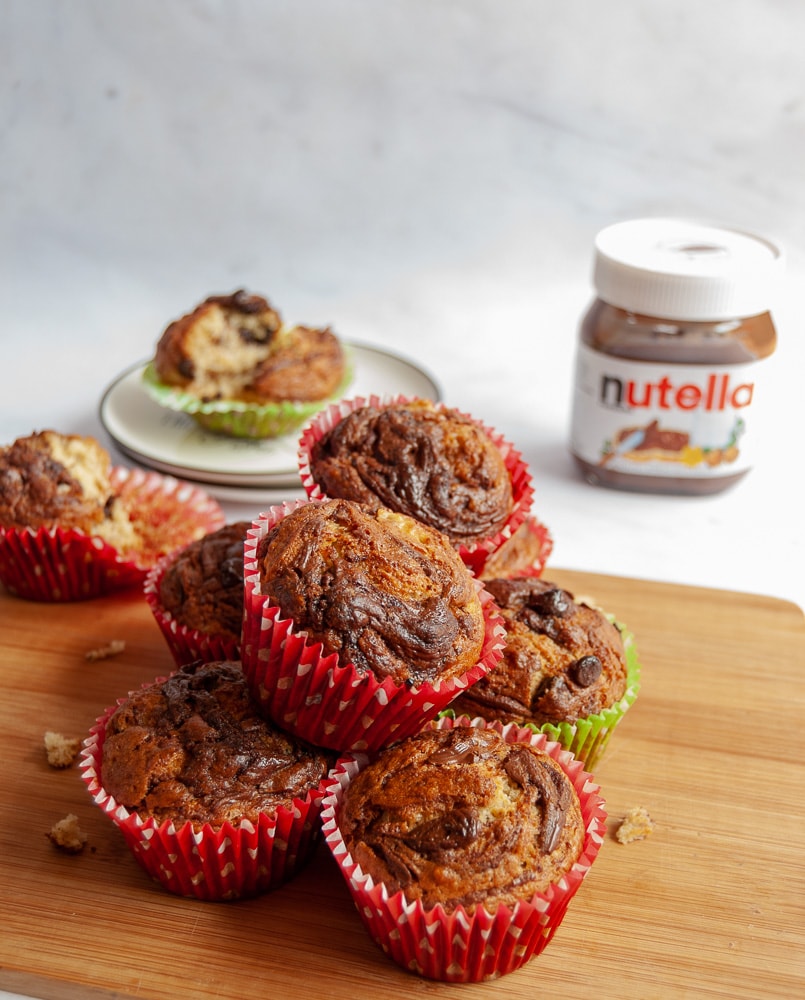chocolate chip banana nutella muffins all piled up on a wooden board. In the background is a plate with a torn open muffin beside a jar of Nutella