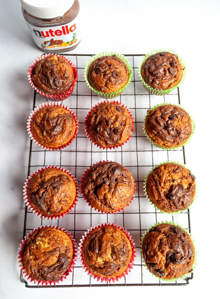 A flat lay photo of twelve chocolate chip banana nutella muffins on a wire rack and white background.