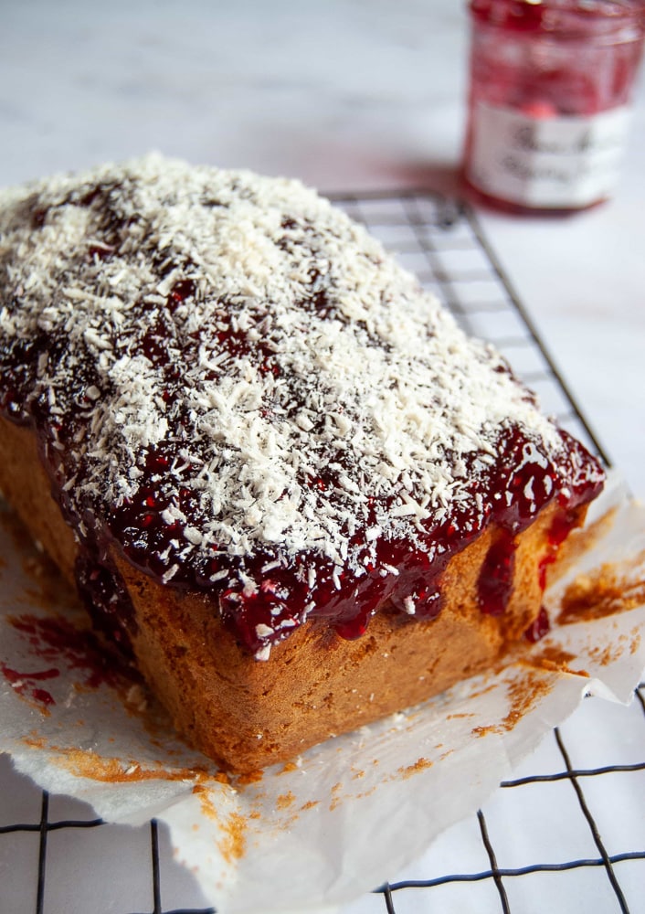 A coconut and raspberry jam loaf cake on a wire rack, with a jar of raspberry jam in the background