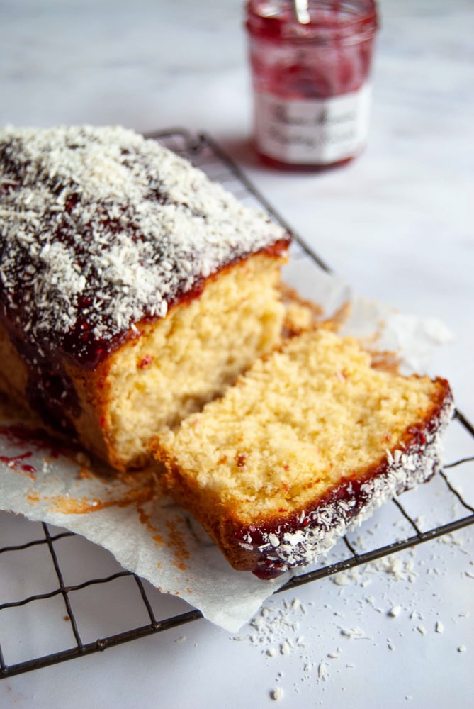 A coconut and raspberry jam loaf cake on a wire rack, with a jar of raspberry jam in the background