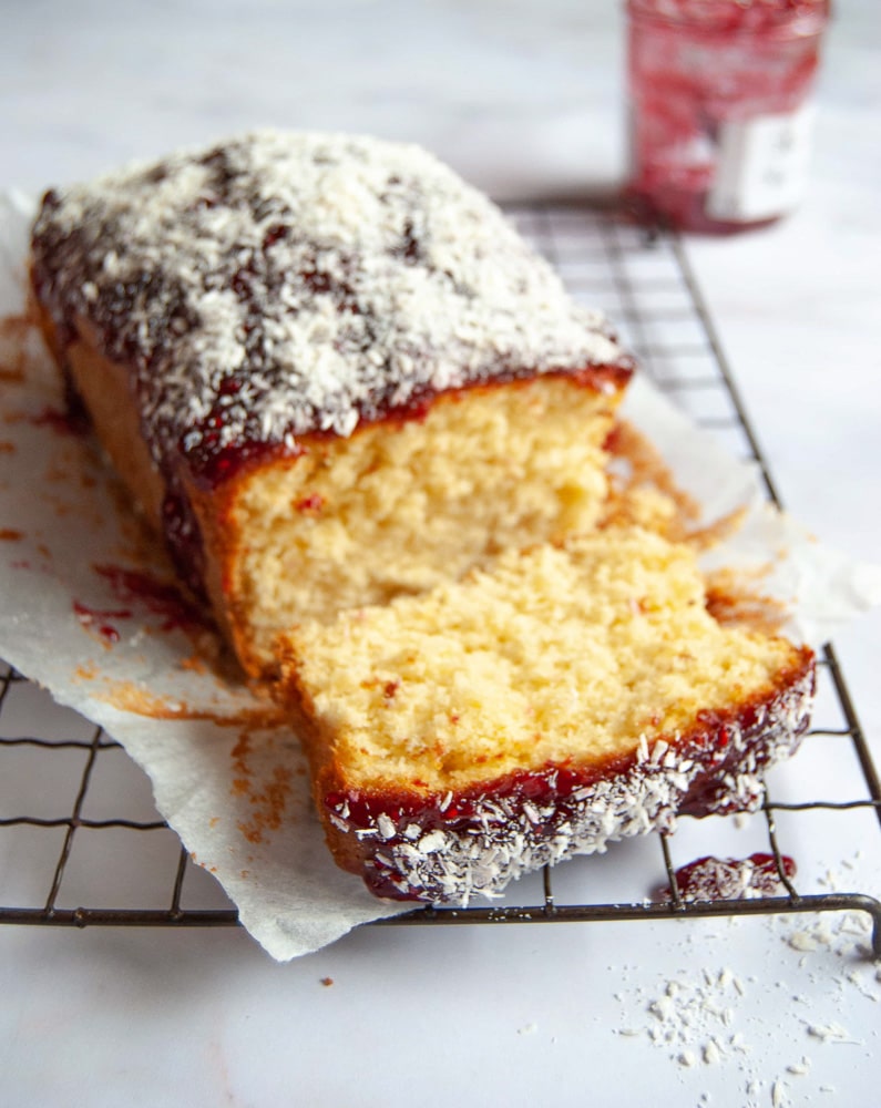 A coconut and raspberry jam loaf cake on a wire rack, with a jar of raspberry jam in the background