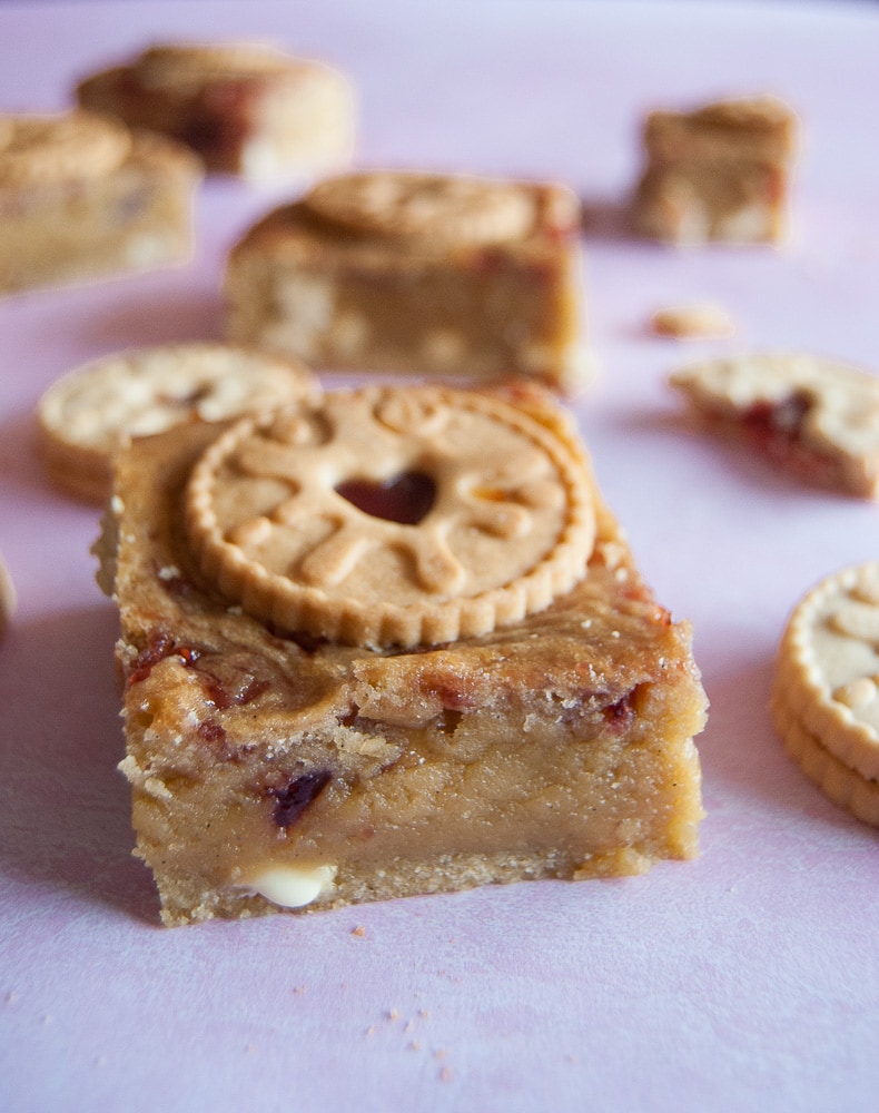 A close up photo of a blondie filled with white chocolate and jam swirls on a light pink background