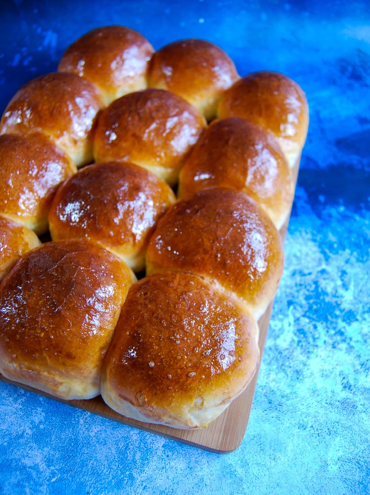 a batch of twelve brioche rolls glazed with egg on a wooden board and blue background.