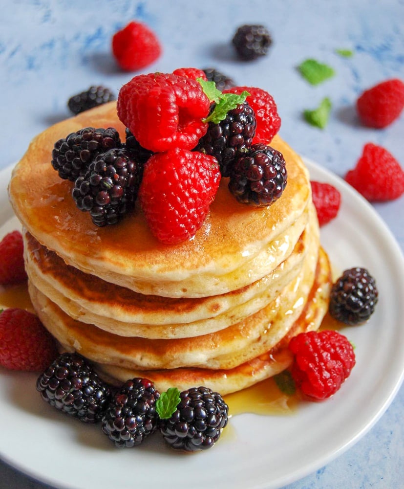 A close up photo of a stack of fluffy American pancakes topped with fresh berries and drizzled with maple syrup.