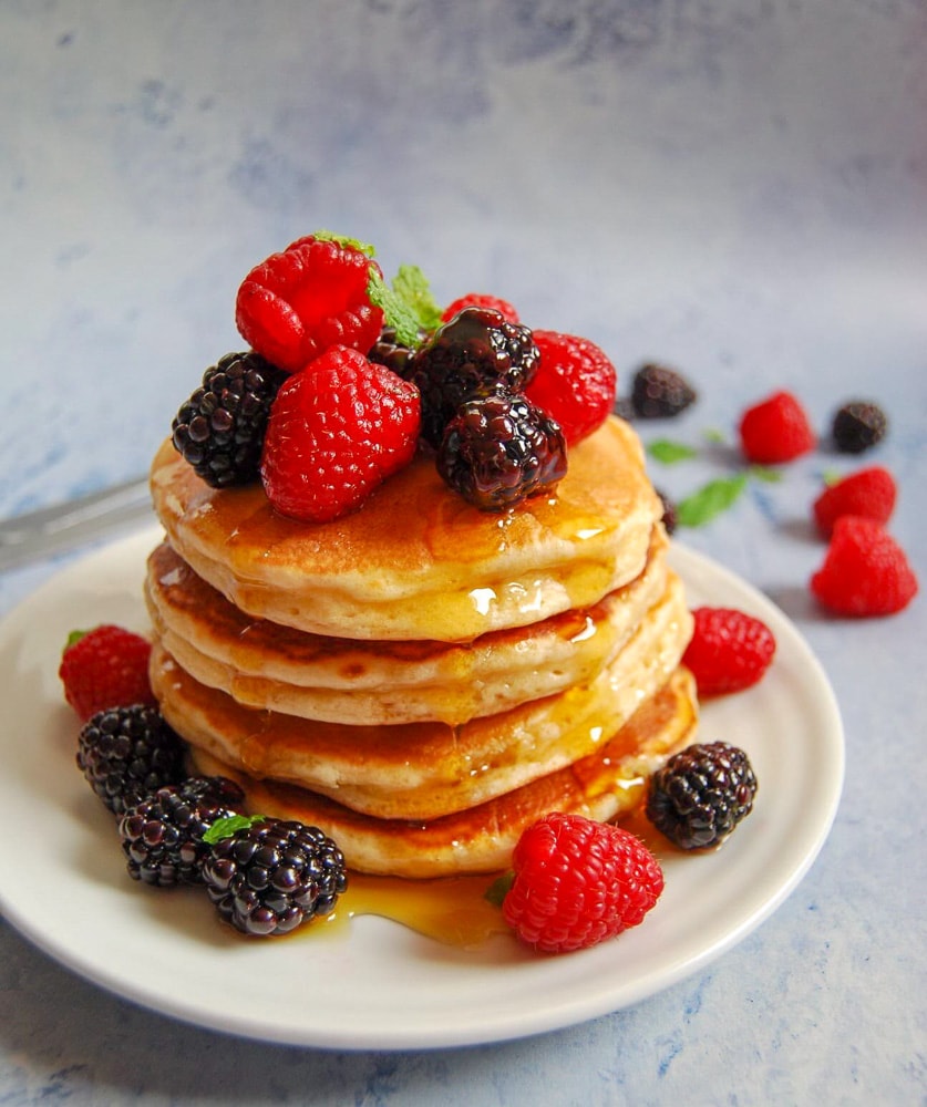 A stack of American style pancakes on a white plate topped with fresh berries and dripping in maple syrup. Background is a mottled light blue.