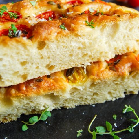 A close up photo of Focaccia bread with cherry tomatoes and fresh thyme. on a black plate. Red and yellow cherry tomates can be seen in the background.