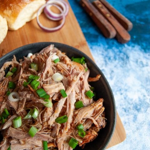 A bowl of slow cooker pulled pork scattered with spring onions. Brioche rolls, red onion slices and cutlery are in the background