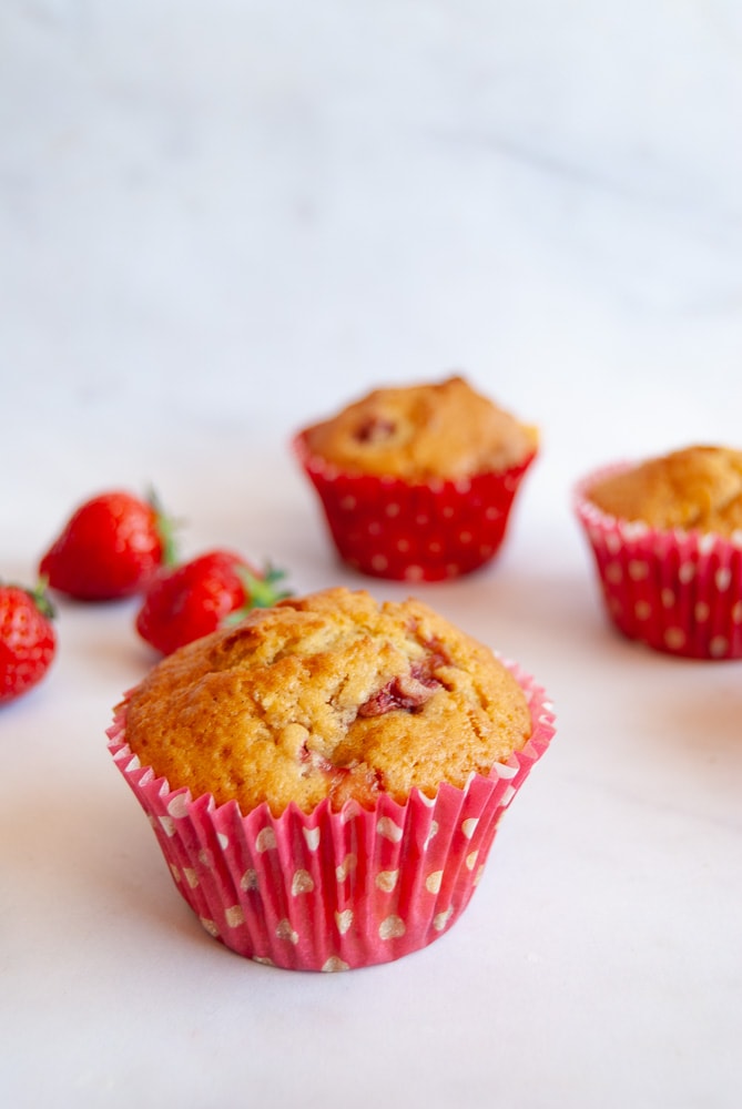 A strawberry muffin with white chocolate chips on a white marbled background. More muffins and fresh strawberries can be seen in the background
