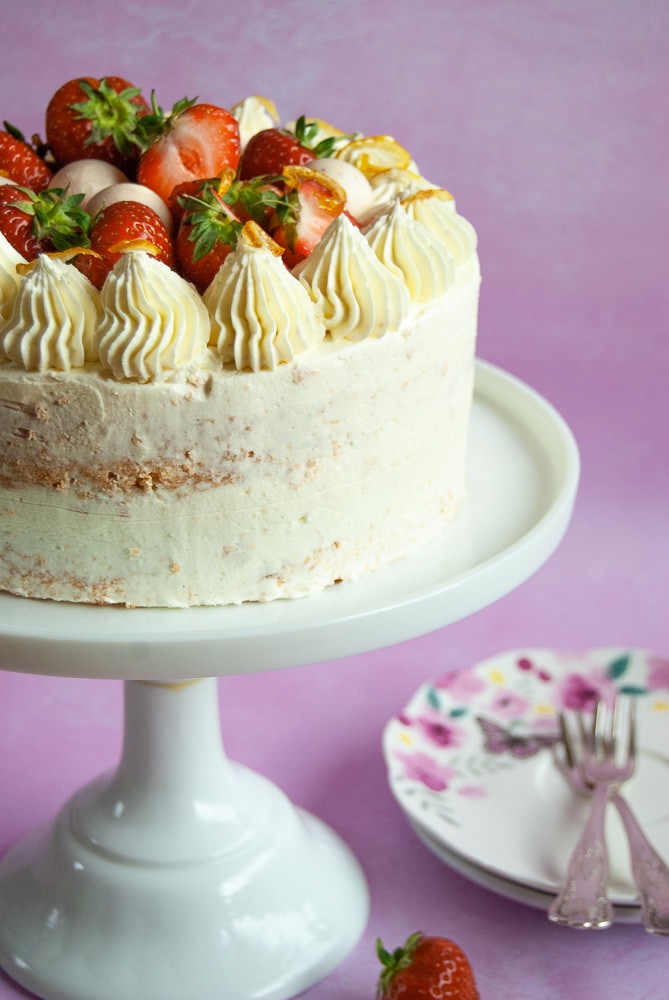 A lemon and elderflower cake with strawberries on a white cake stand. The cake has been decorated with buttercream swirls and topped with fresh strawberries, Lindor truffles and candied lemon peel.