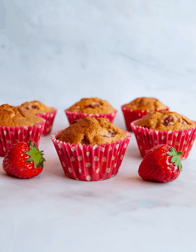 six strawberry muffins with white chocolate chips in pink spotted muffin cases on a white backdrop and two fresh strawberries.