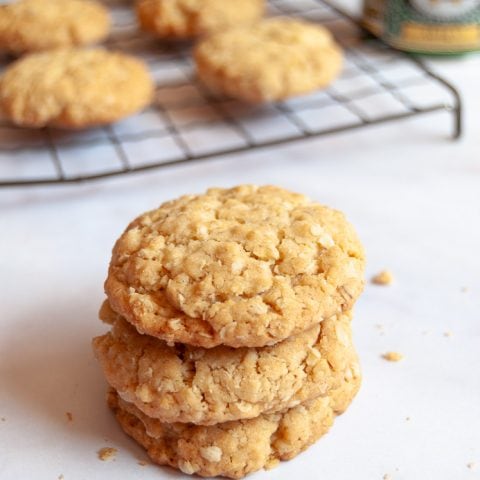 Three oat biscuits stacked on top of each other on a white background. A wire rack with more oat biscuits and a tin of golden syrup can be seen in the background.