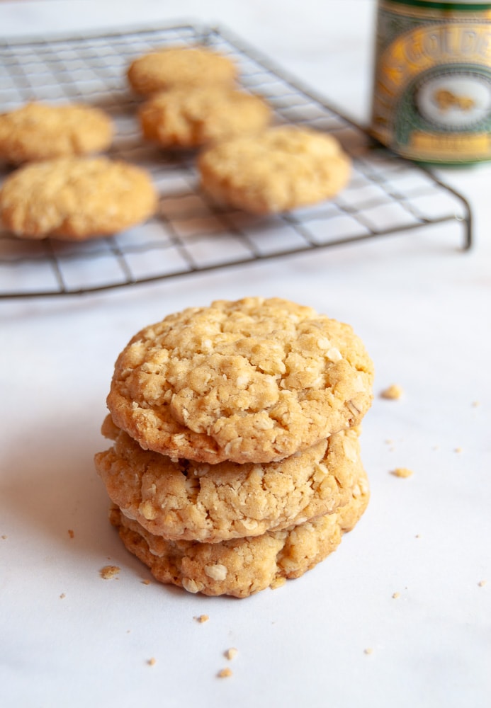 Three oat biscuits stacked on top of each other on a white background. A wire rack with more oat biscuits and a tin of golden syrup can be seen in the background.