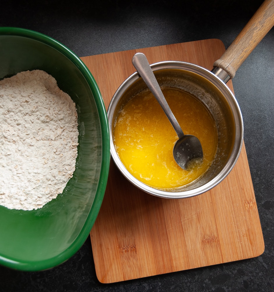 A pan of melted butter and a green bowl of flour and oats in a wooden board 