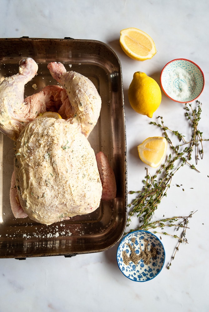 A photo of an uncooked raw chicken in a roasting tin covered in a herb butter.