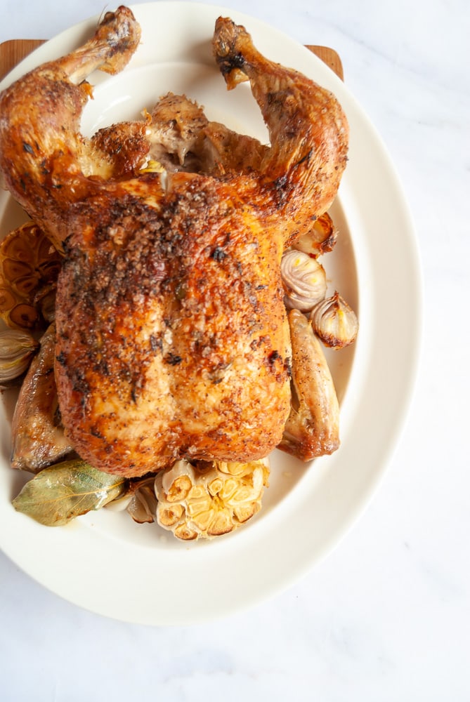 A picture of a cooked Chicken on a white oval plate with roasted shallots, garlic bulbs and bay leaves.