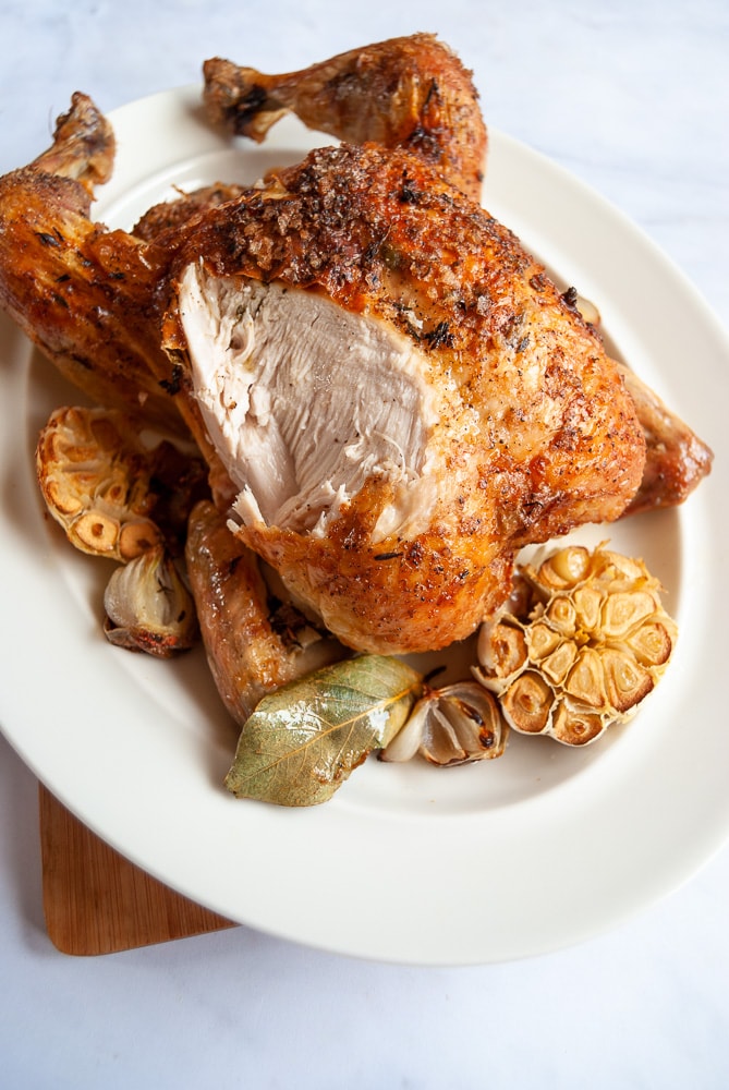 a photo of a cooked roast chicken on a white oval plate with a roasted garlic bulb cut in half and a bay leaf. The chicken has been carved to reveal the breast meat.