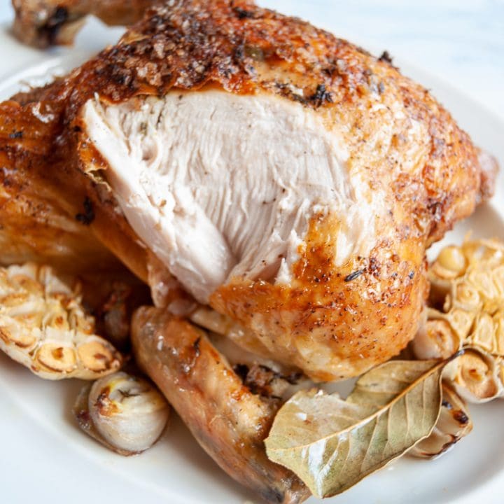 A close up photo of a cooked roast chicken with halved garlic bulbs, shallots and a bay leaf. The chicken has been carved to show the breast meat.