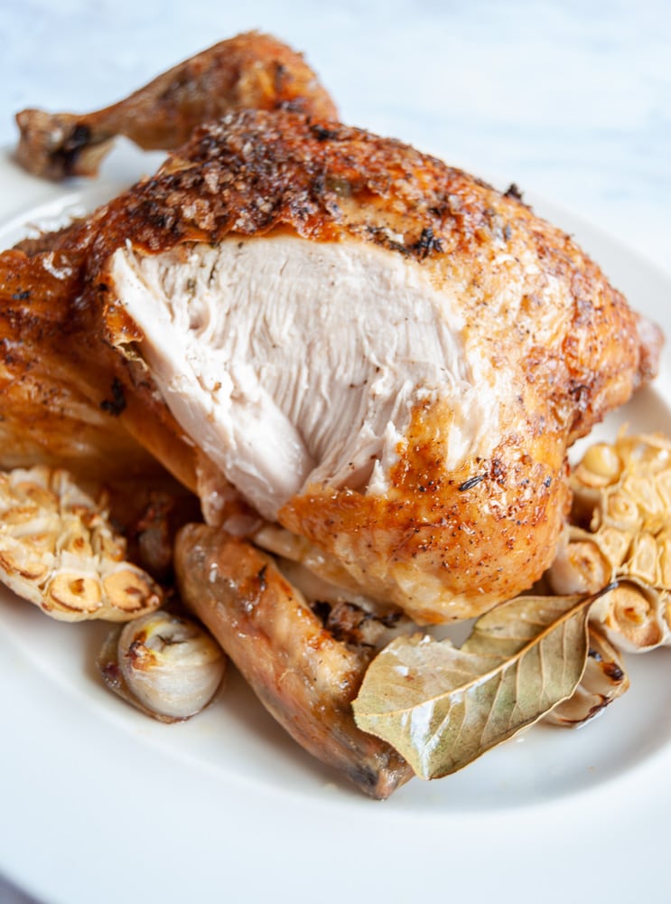 A close up image of a roast chicken with halved garlic bulbs, shallots and a bay leaf. 