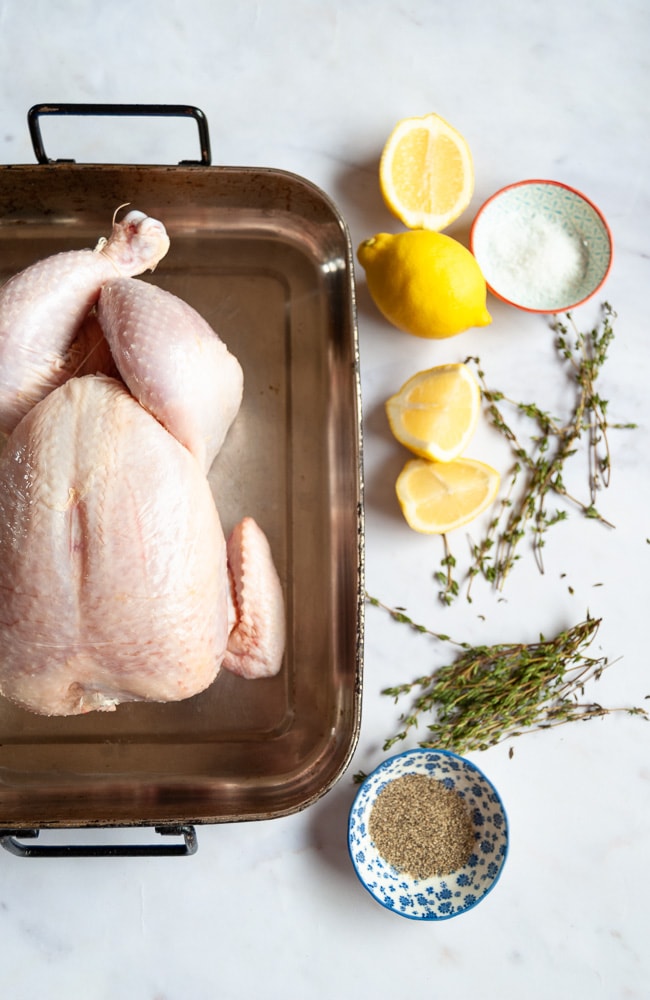 A photograph of an uncooked whole chicken in a roasting tin. Lemons, a pot of sea salt and fresh thyme is beside the chicken.