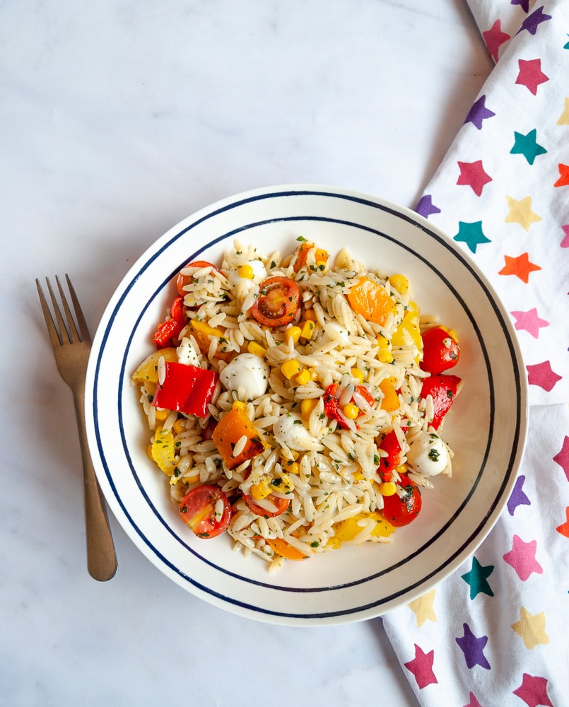 A flat lay photo of a bowl of roasted pepper orzo salad with cherry tomatoes, mozzarella pearls, sweetcorn and fresh chopped herbs on a white background.