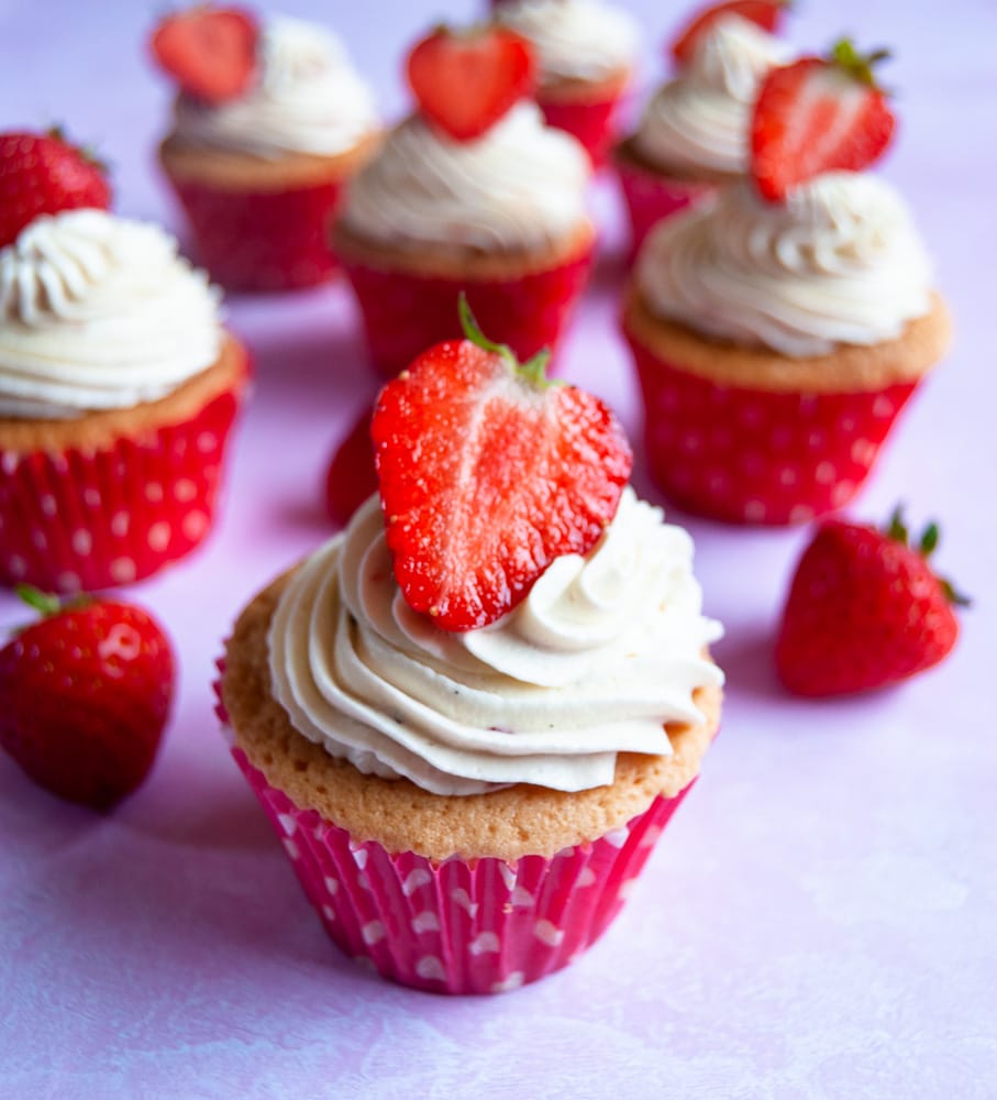 A close up photo of a vanilla cupcake decorated with a fresh whipped cream swirl and topped with a strawberry. More cupcakes can be seen in the background.