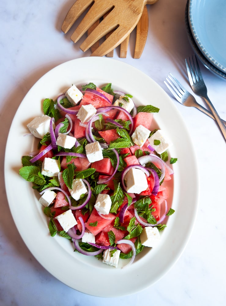 A salad with watermelon chunks, feta cheese, red onion slices and fresh mint leaves on a white platter