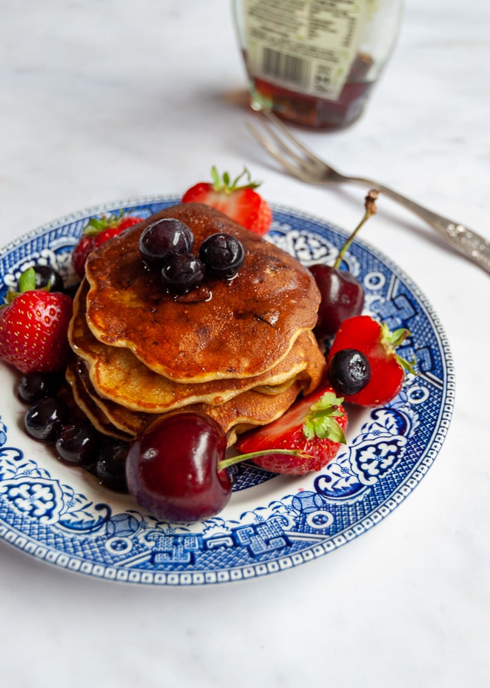 sugar free banana and blueberry pancakes drizzled with maple syrup and topped with fresh fruit on a blue willow pattern plate