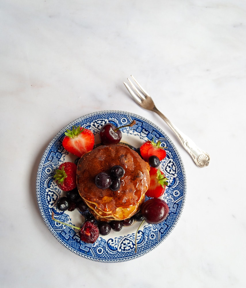 A flat lay photo of a stack of sugar free banana and blueberry pancakes drizzled with maple syrup and topped with cherries, strawberries and blueberries on a blue willow pattern plate.
