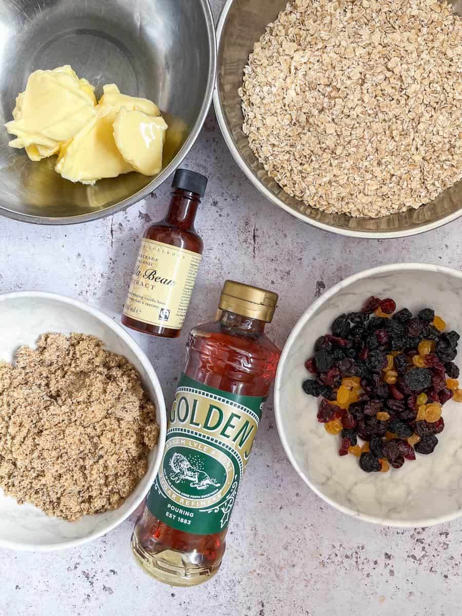 A silver bowl of butter, a silver bowl of rolled oats, a bottle of vanilla extract, a bottle of golden syrup, a bowl of brown sugar and a bowl of dried berries.