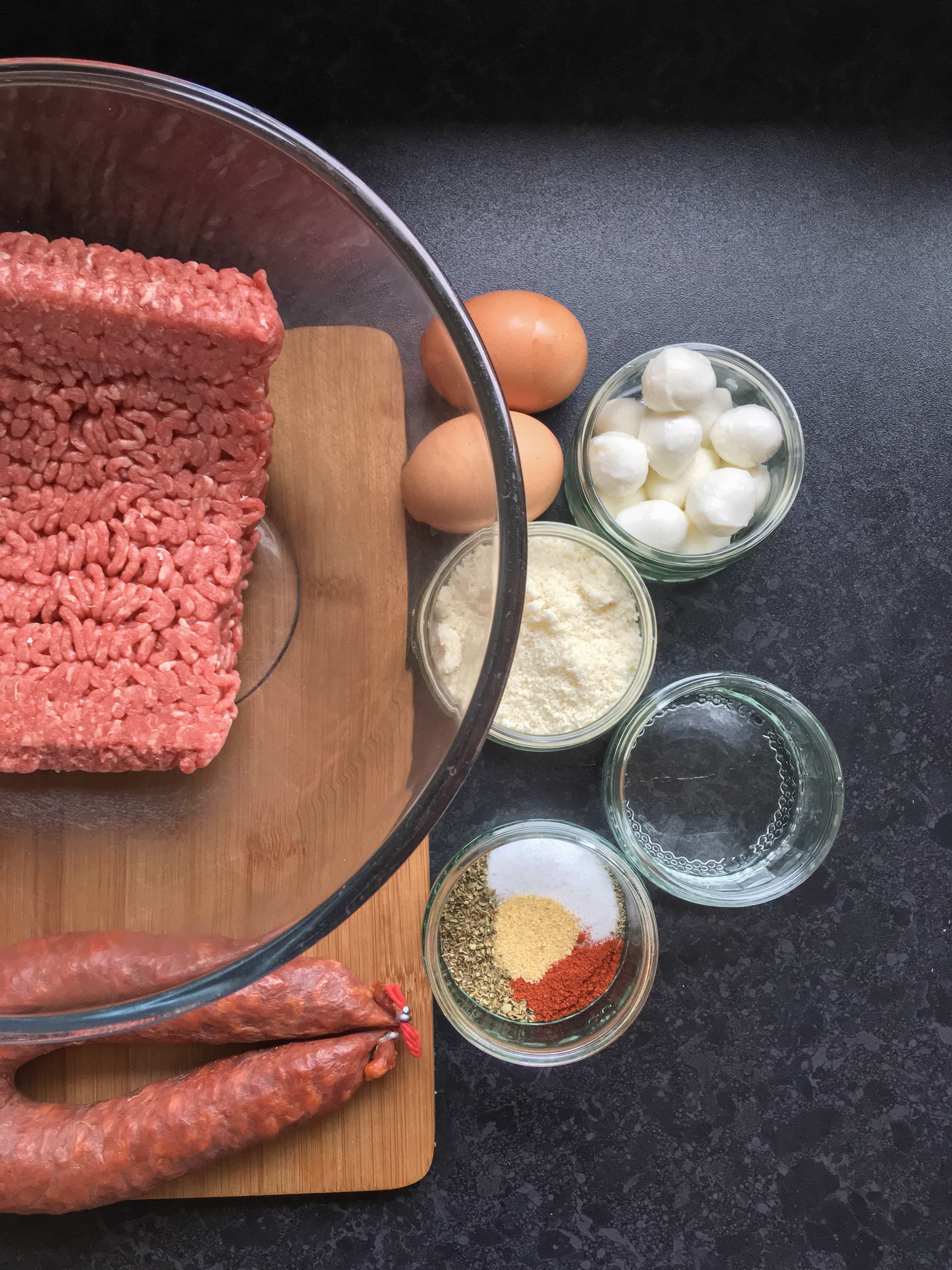 a large pyrex bowl of minced beef, a chorizo sauce, two eggs and small glass dishes of mozzarella pearls, parmesan cheese and ground spices.