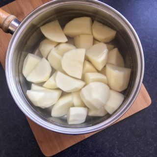 A saucepan of water and peeled, quartered potatoes on a wooden chopping board.