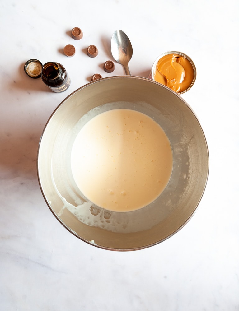 a large silver bowl filled with cream, a tin of caramel, a spoon, a bottle of vanilla extract and Rolo candies on a white background.