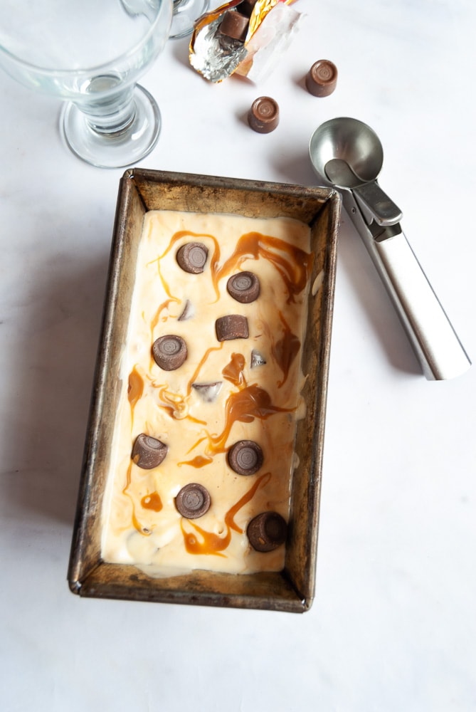 A flat lay photo of a loaf tin with salted caramel ice cream studded with Rolo candies.