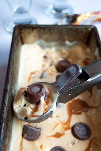 A close up photo of a scoop of no churn rolo ice cream with salted caramel swirls. Ice cream Sundae glasses and an opened packet of Rolo candies can be just seen in the background.