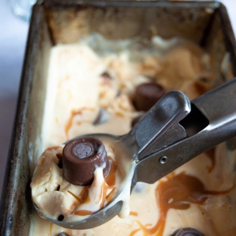 A close up photo of a scoop of no churn rolo ice cream with salted caramel swirls. Ice cream Sundae glasses and an opened packet of Rolo candies can be just seen in the background.