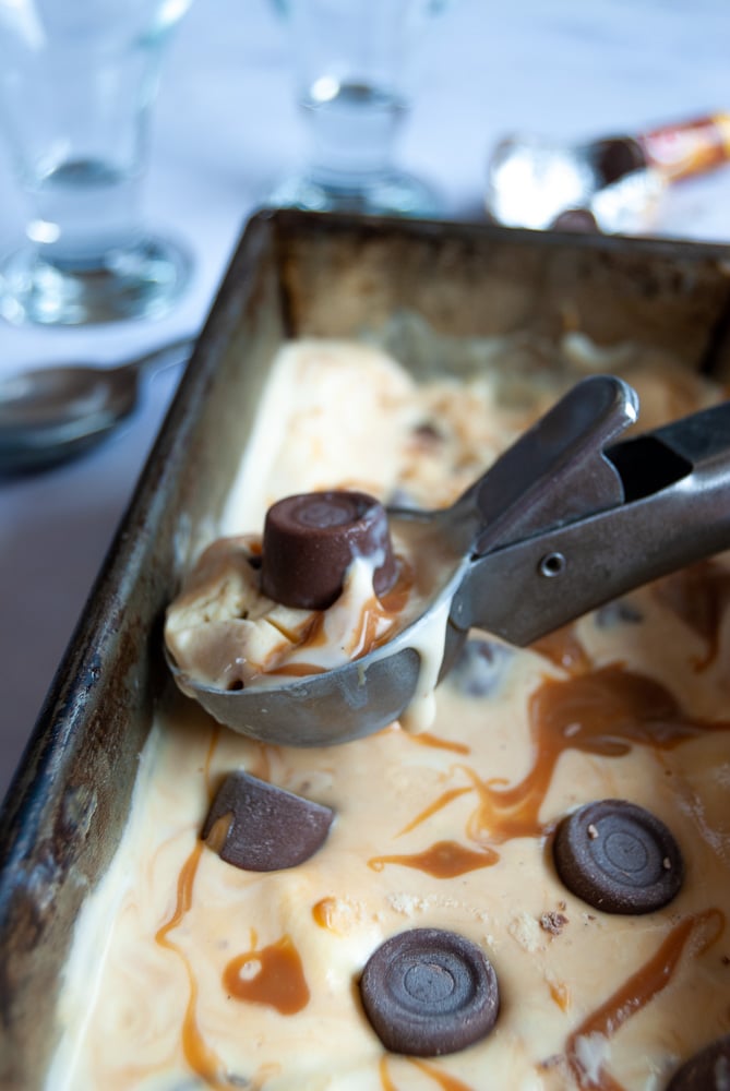 A loaf tin with no churn salted caramel ice cream. A silver ice cream scoop with a scoop of the ice cream sits on top of the tin.