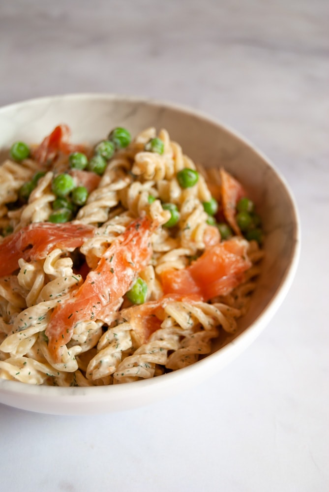 A white bowl of sweet chilli pasta with smoked salmon slices, peas and chopped dill.