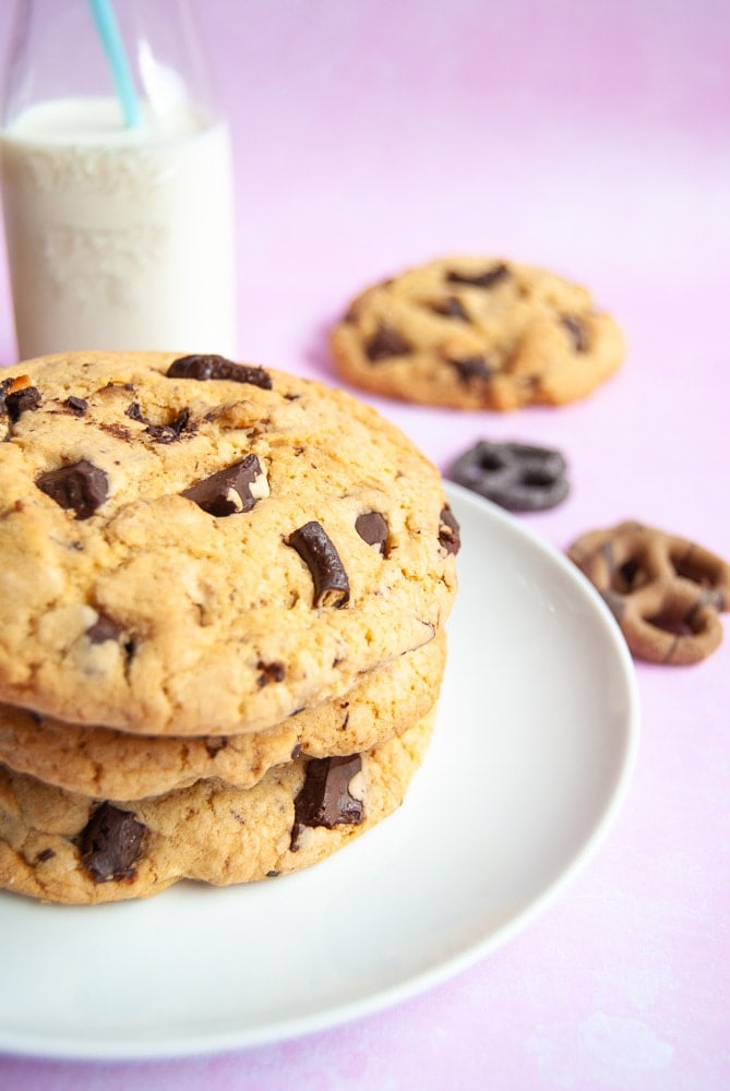 A white plate of chocolate pretzel cookies on a pink backdrop. A jug of milk and chocolate pretzels are also in the photo