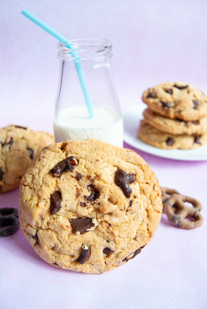 A large chocolate pretzel cookie resting on a mini bottle of milk. More cookies on a white plate are in the background. 