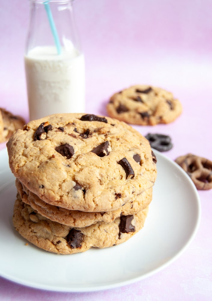 A stack of chocolate pretzel cookies on a white plate. A mini bottle of milk and more cookies and chocolate pretzels are in the background. 