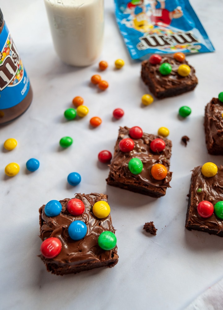 Chocolate Brownies topped with M&M spread and M&M candies on a white backdrop. A jar of M&M spread, a bottle of milk and a spilled packet of M&Ms are in the background